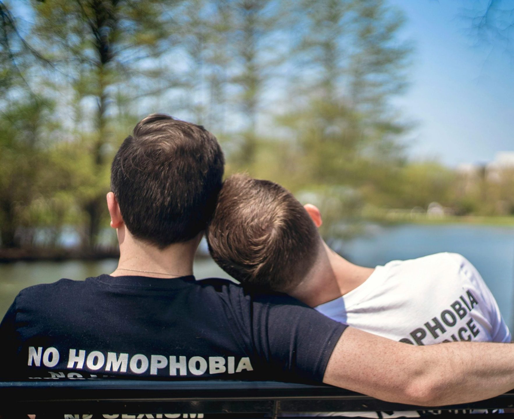 Two men sitting on a park bench by a lake, embracing love with unity and pride.